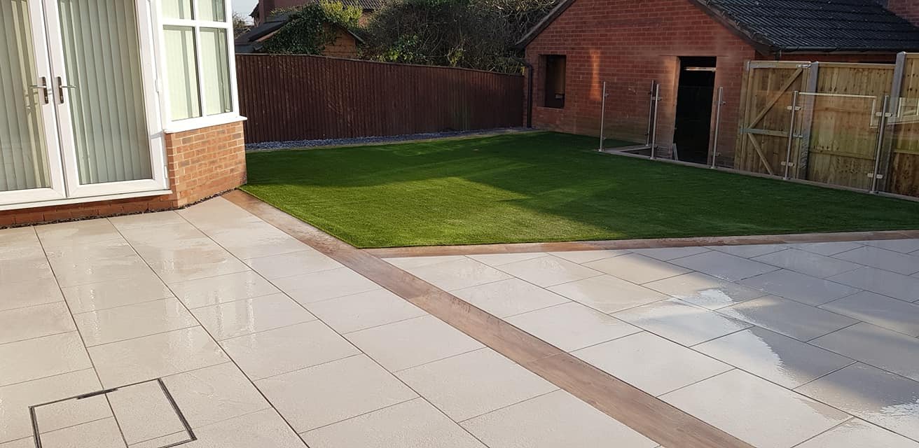Backyard with beige tiled patio, green artificial grass lawn, wooden fencing, and a brick outbuilding.