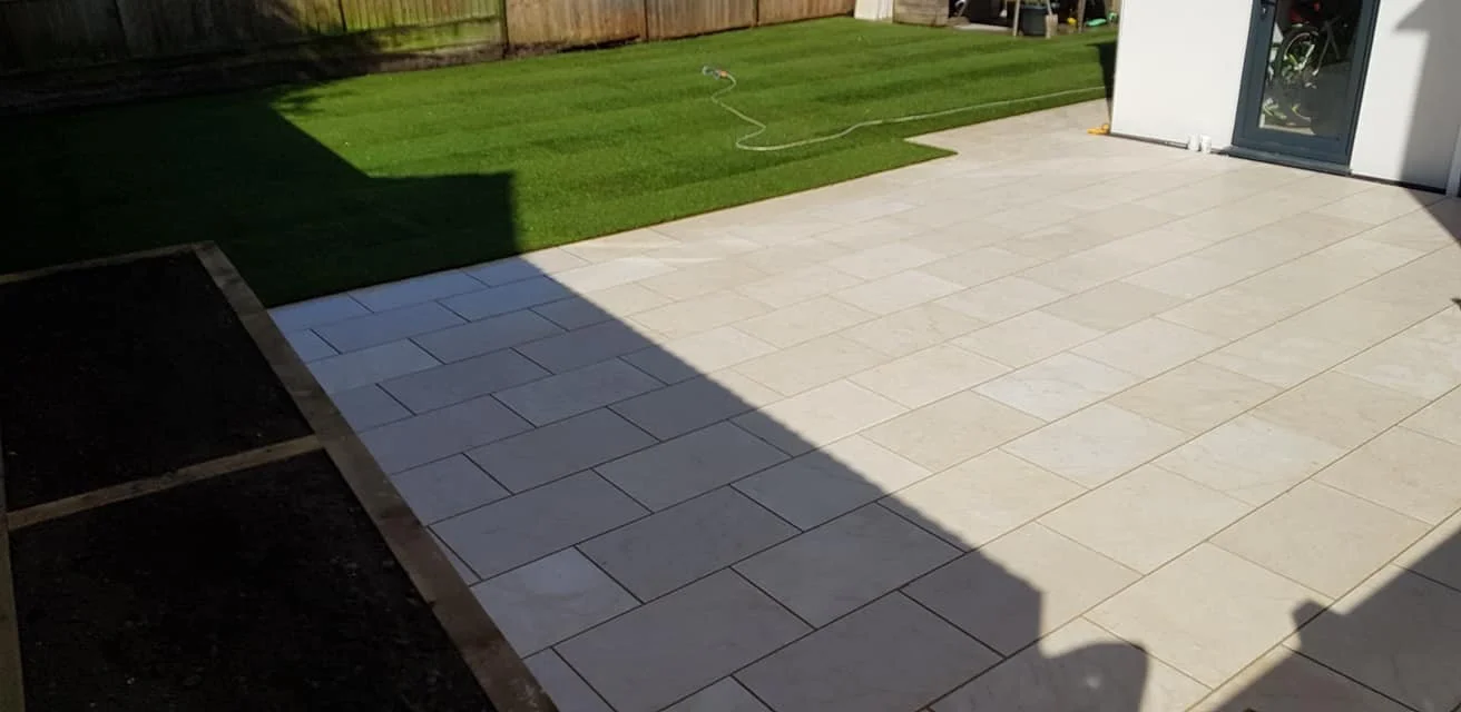 A backyard patio with light-colored tile flooring, adjacent to a grassy lawn. Part of the patio is in shadow. There's a garden hose on the grass and a white building with glass doors on the right.