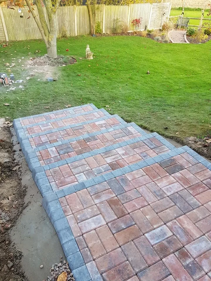 Partially completed brick patio in a backyard with a grassy lawn, trees, and a wooden fence.