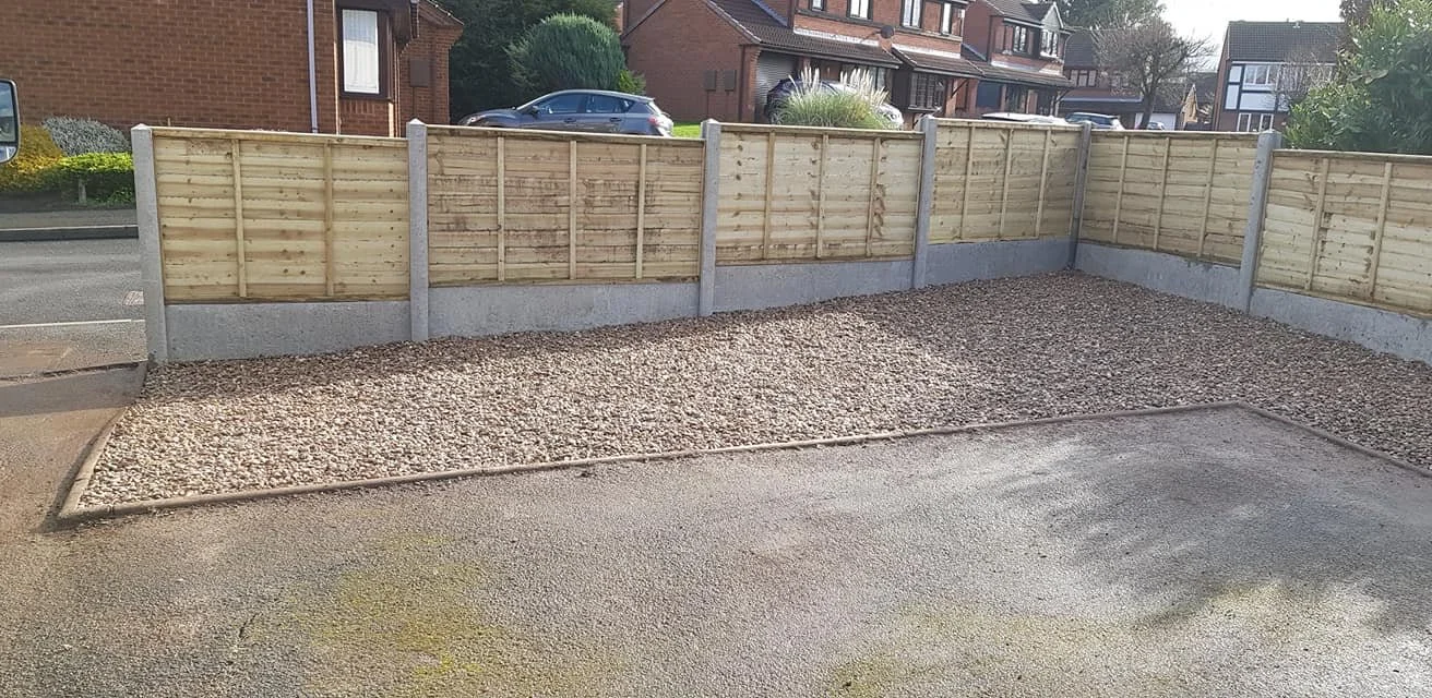 A fenced parking area with gravel ground and a curb separating it from an asphalt road.