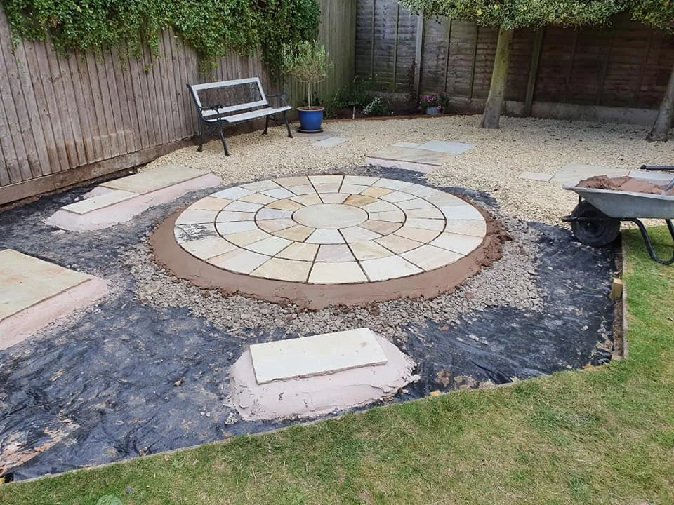 Garden backyard with a partially completed circular stone patio, a black and white bench, a potted plant, and a wheelbarrow on the grass.