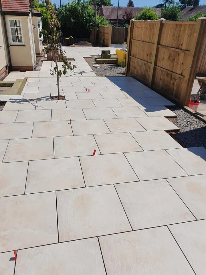 A backyard patio with newly installed beige stone tiles, a small tree planted in the middle, a wooden fence on the right, and a gravel area with construction materials in the background.