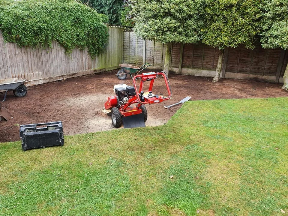 Backyard with a lawn and a dug-up dirt patch for landscaping, including a red machine, a wheelbarrow, and a level tool.