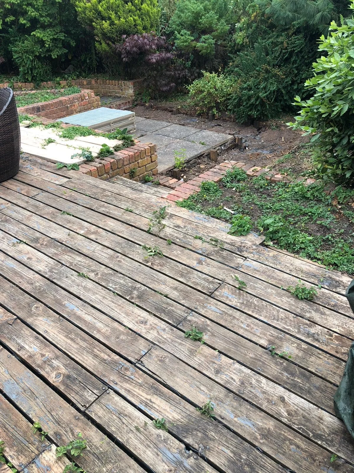 Backyard patio with a wooden deck, overgrown plants, and a small brick-paved area with some uneven tiles and a garden bed.