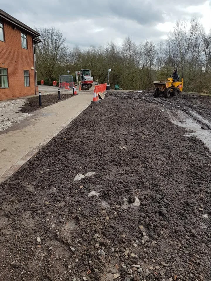 Construction workers are working on a sidewalk and pathway, with a bulldozer and a compact excavator on site. The area has exposed dirt, and orange safety cones are placed along the pathway. A brick building is visible on the left, and trees are in t