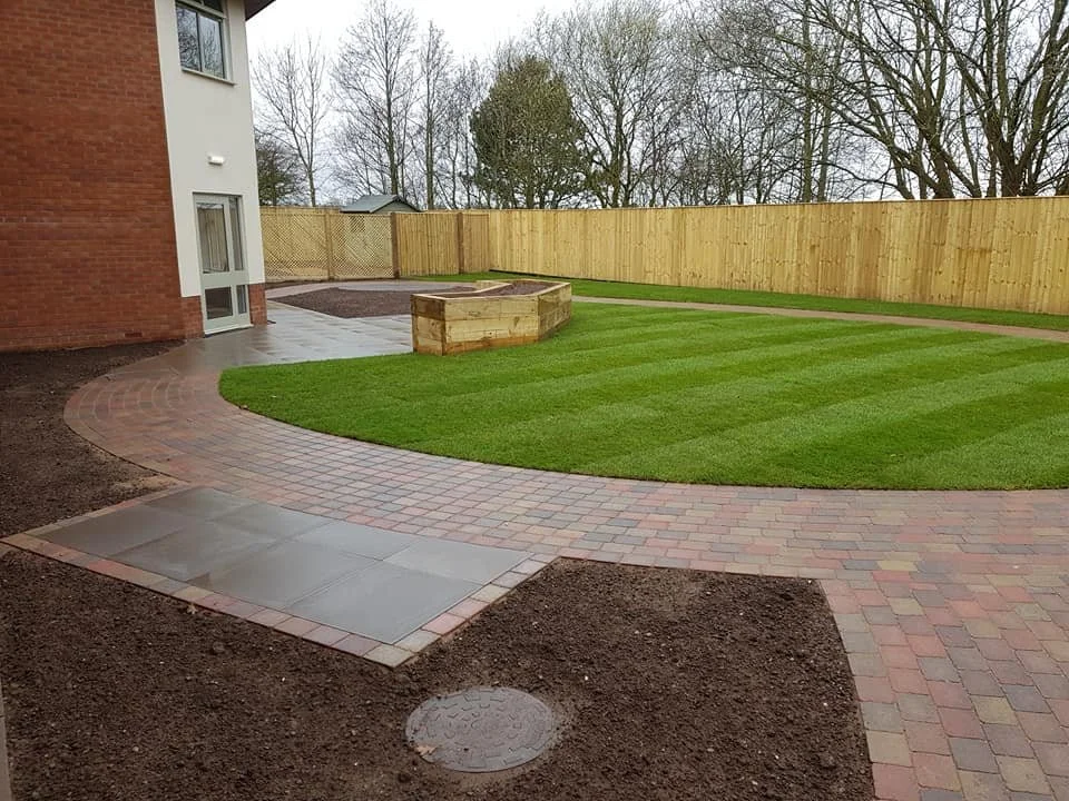 Newly landscaped backyard with a green lawn, brick walkway, and wooden planter box, enclosed by a wooden fence, in a suburban setting.