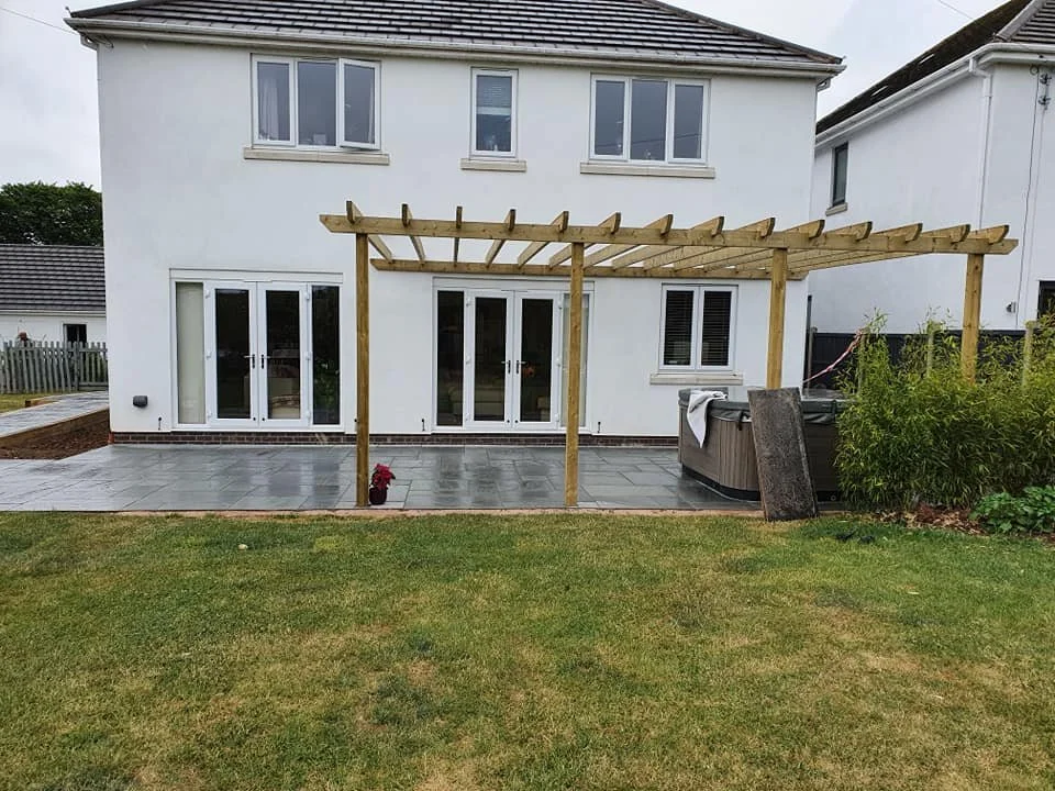 Backyard patio area with a wooden pergola frame, part of a two-story white house, sliding glass doors, a hot tub with a cover, and some shrubs on the right side.