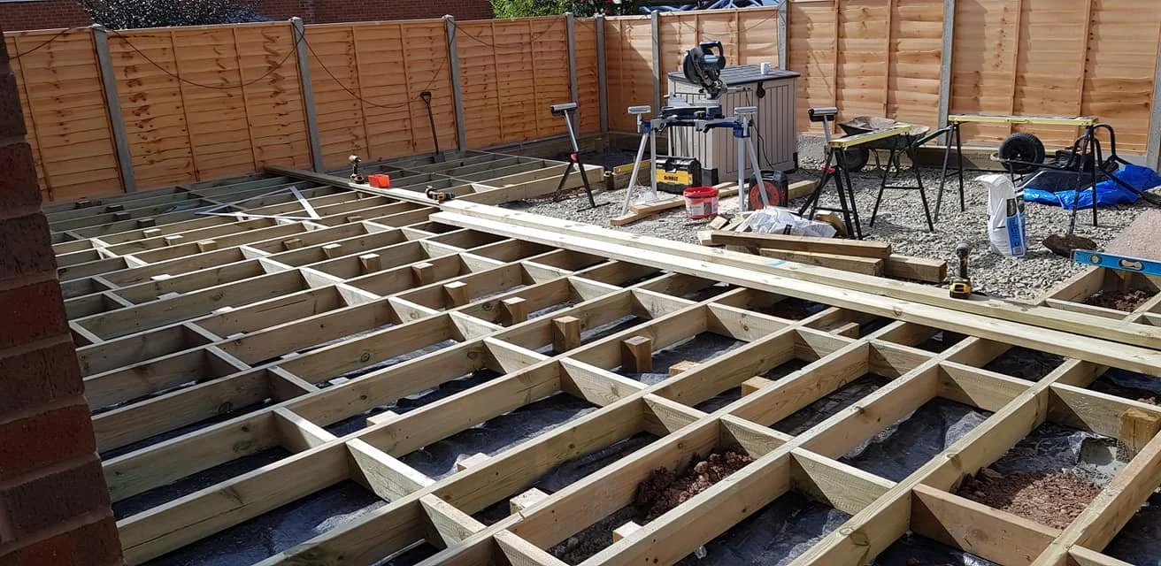 A backyard deck under construction with a wooden frame, construction tools, and equipment, enclosed by a wooden fence.
