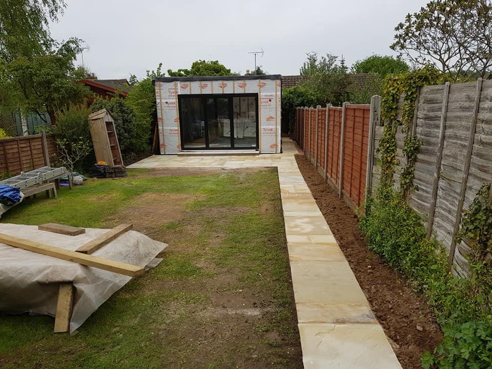 Backyard garden area with a newly laid stone pathway leading to a small building under construction, with wooden fences and some plants and tools visible.