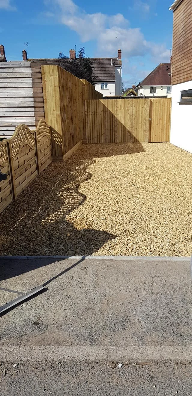 Newly installed wooden privacy fence in a backyard with gravel ground and neighboring houses in the background under a partly cloudy sky.