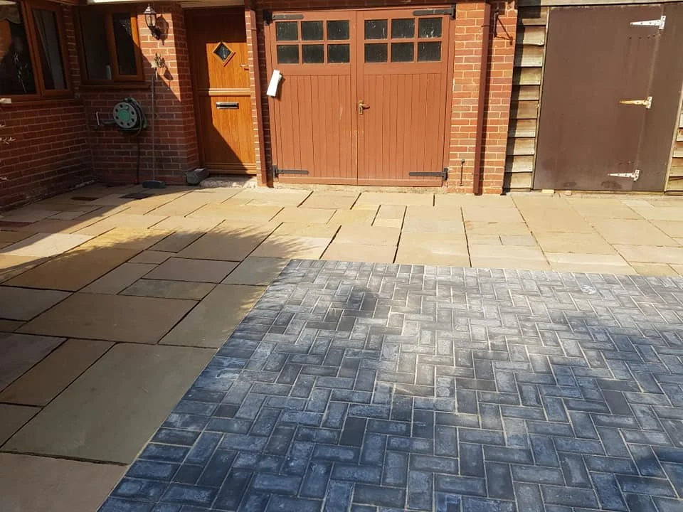 A paved backyard with beige and gray bricks, a brick house with a wooden door, a garage door, and a wooden side gate.