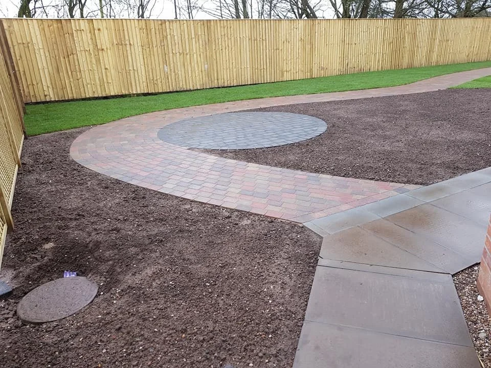 A backyard with a curved brick pathway, a round concrete stepping stone, and a wooden privacy fence. The yard has areas of green grass and brown soil.