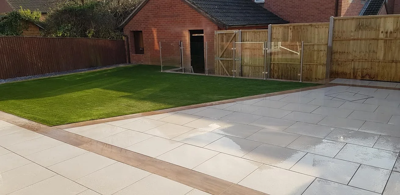 Backyard with a new green lawn, a paved patio area, and a wooden fence fence, with a brick house in the background.