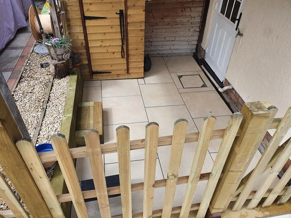Small fenced patio area with beige tiles, a wooden storage shed, a white door, and gravel border with a flower pot.