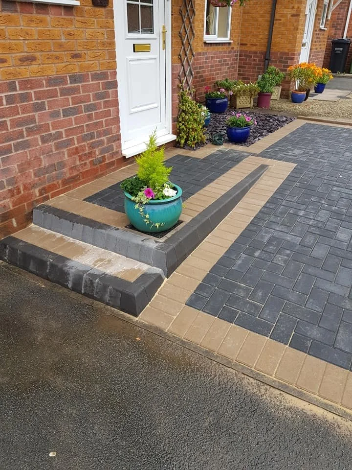 Entrance to a brick house with white door and several potted plants on the front porch and driveway, featuring a patterned brick and concrete pavement.