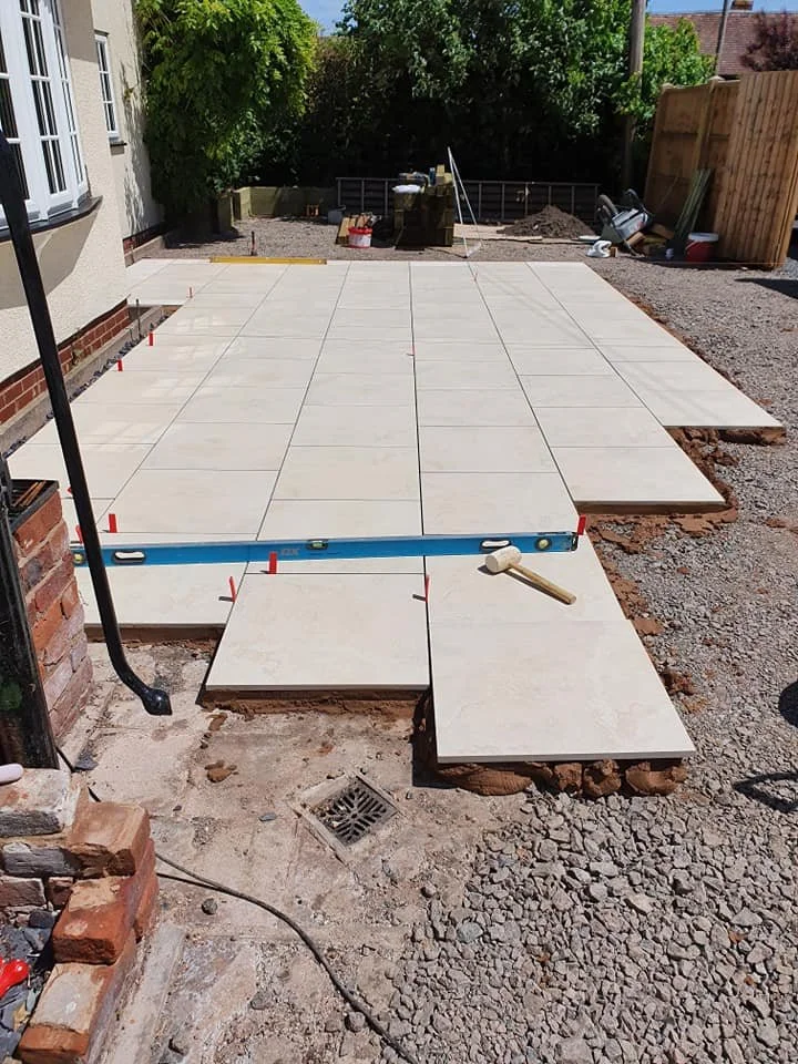 Outdoor patio construction with large beige tiles being laid on the ground next to a house, with some tiles already installed and others waiting to be placed. Tools and construction materials are visible, along with a level on the ground.