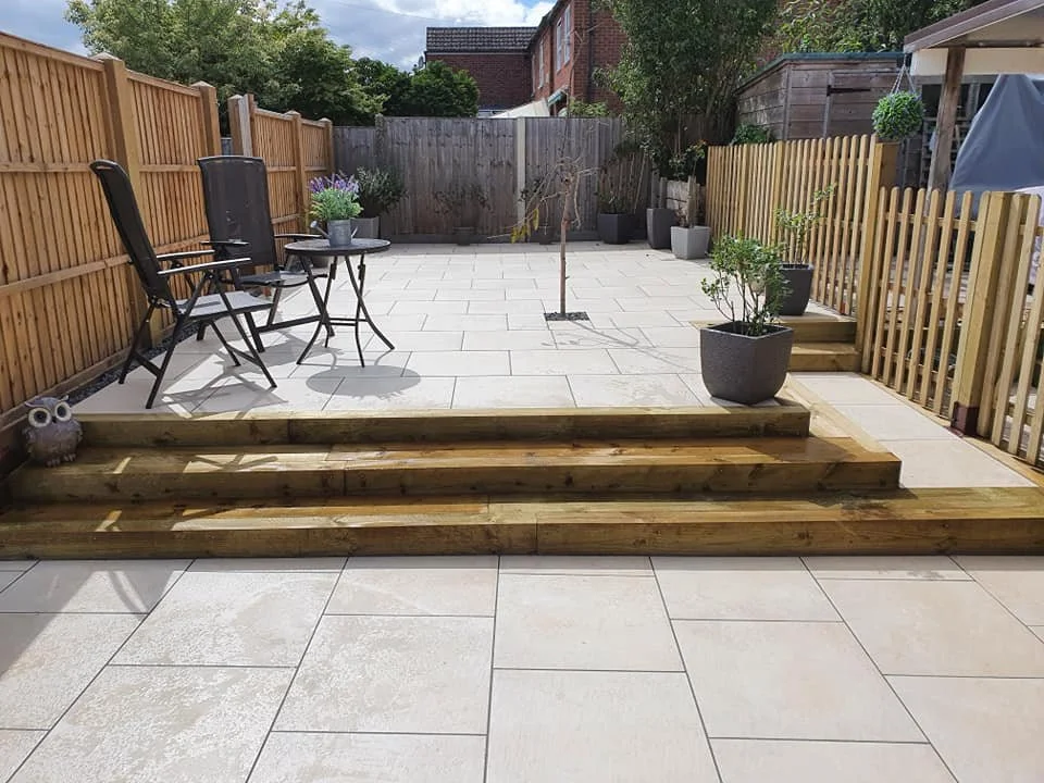 A backyard patio with large beige tiles, a wooden step, potted plants, a small tree, outdoor chairs and a fence, under a partly cloudy sky.