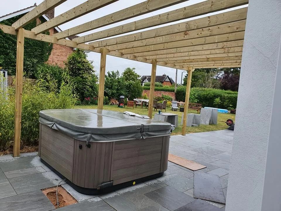 Backyard patio with a hot tub, unfinished pergola, outdoor furniture, and a garden with trees and shrubs.