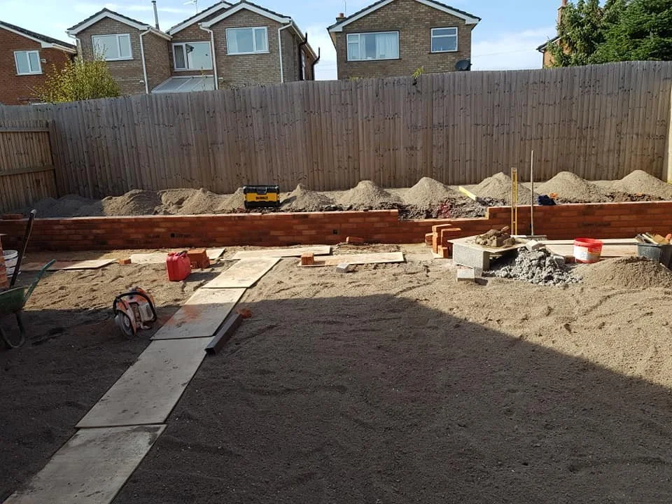 Backyard under construction with brick wall in progress, dirt ground, construction tools, and supplies, surrounded by a wooden fence and residential houses in the background.
