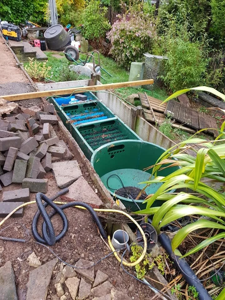 Backyard construction site with a green water filter or pond system being installed, surrounded by bricks, pipes, and tools.