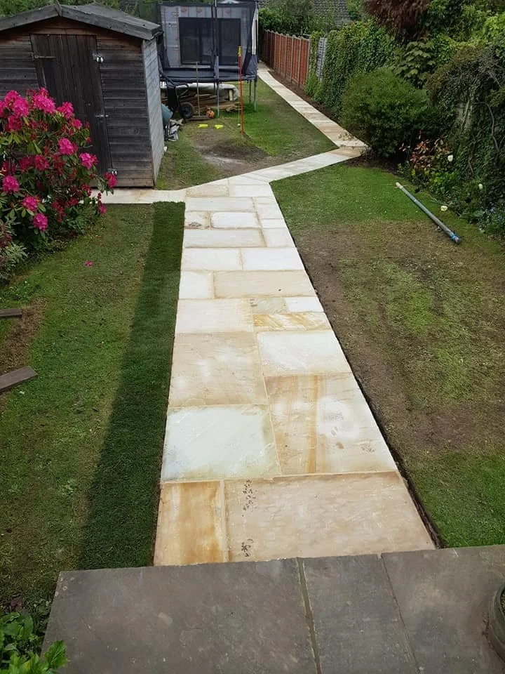 Backyard garden pathway with beige stone tiles, bordered by grass and a shed, leading to a trampoline in the corner, surrounded by bushes and a wooden fence.