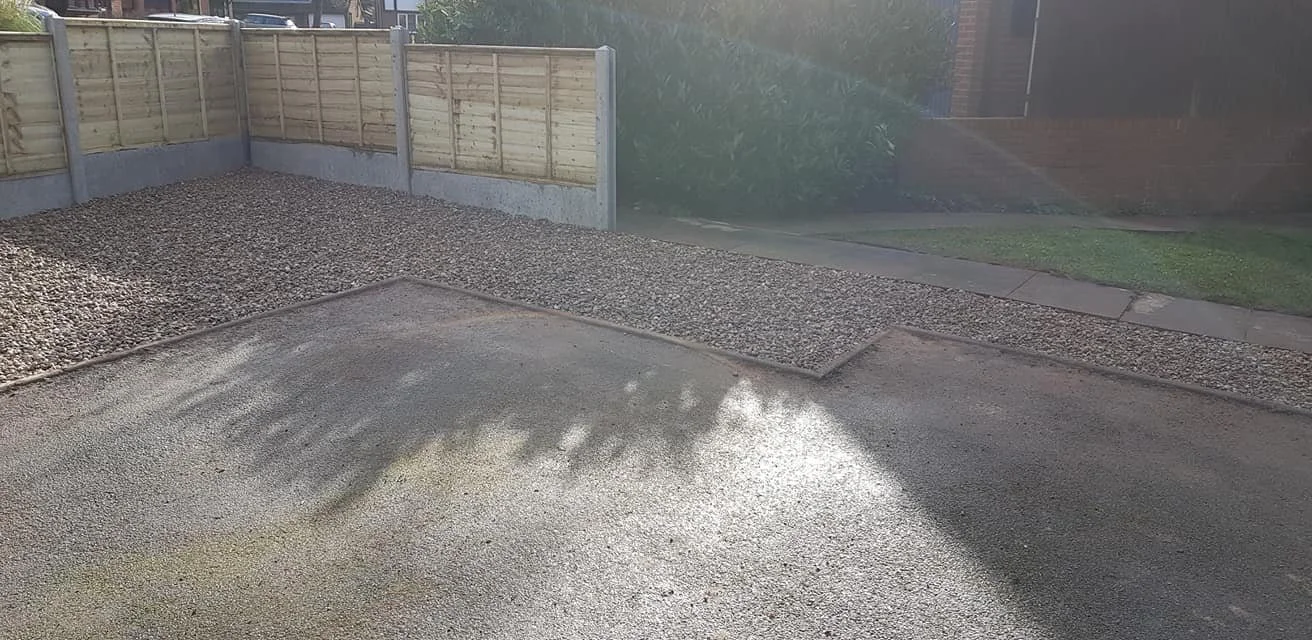 A driveway with fresh asphalt, a gravel area, and a wooden fence in the background.