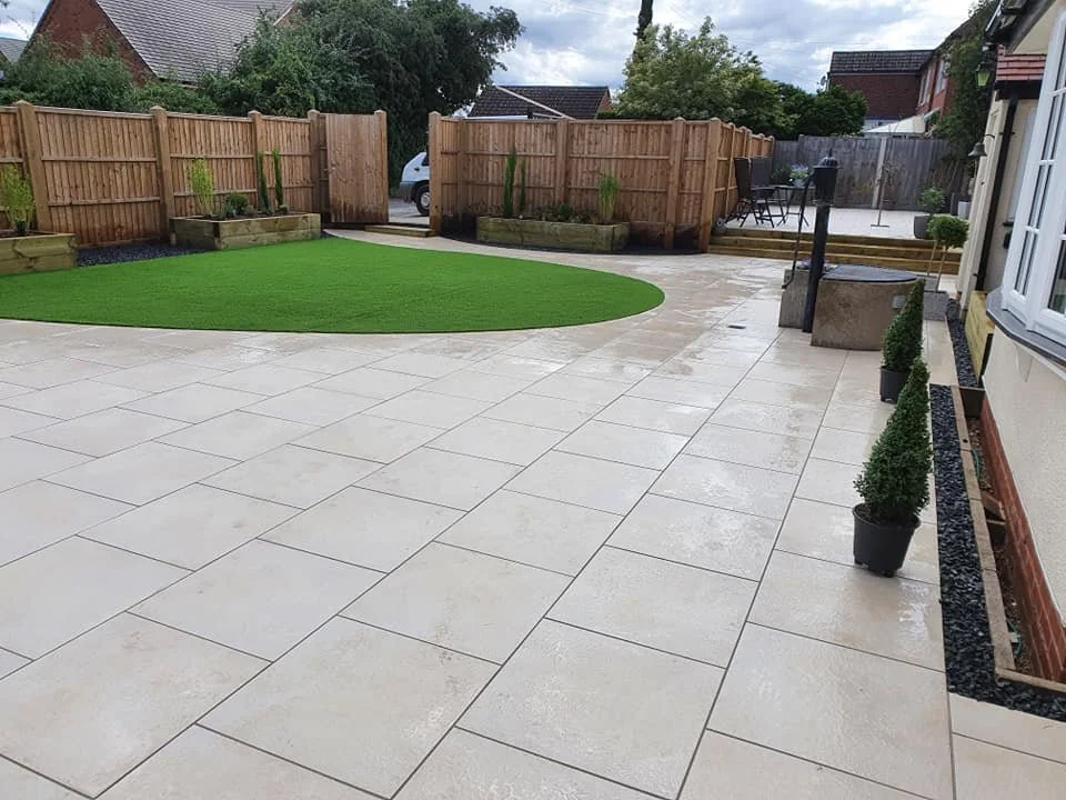A backyard patio with tiled flooring, a section of artificial grass, potted plants, and a wooden deck with outdoor furniture, enclosed by a wooden fence.