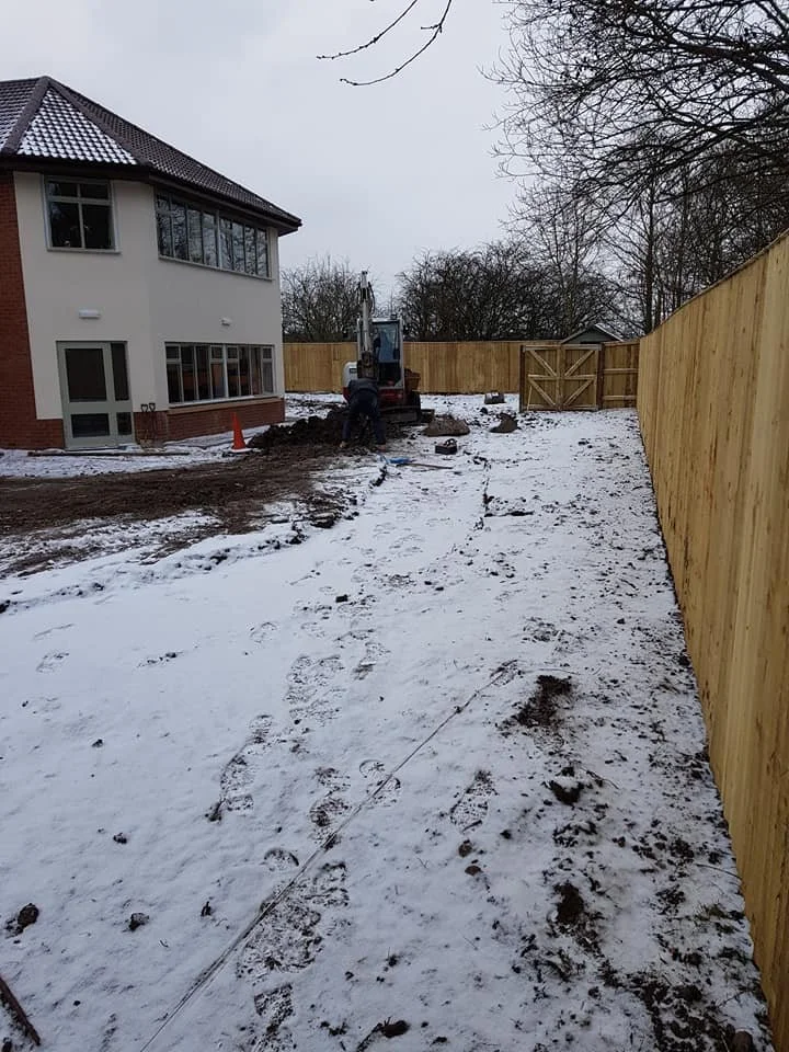 Construction site in a snowy backyard where workers are installing a new fence, with a small excavator and a partially built wooden gate.