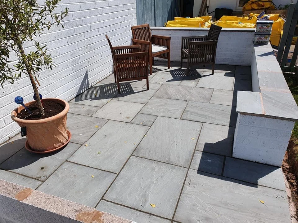 Small outdoor patio with three wooden chairs, a potted plant, and some yellow bags or tarps in the background.