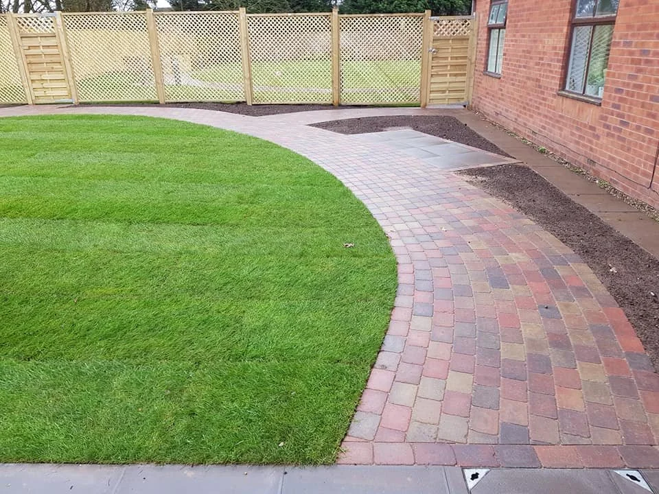 A backyard with a green lawn, a curved brick pathway, soil beds, a wooden fence, and a brick house wall with windows.