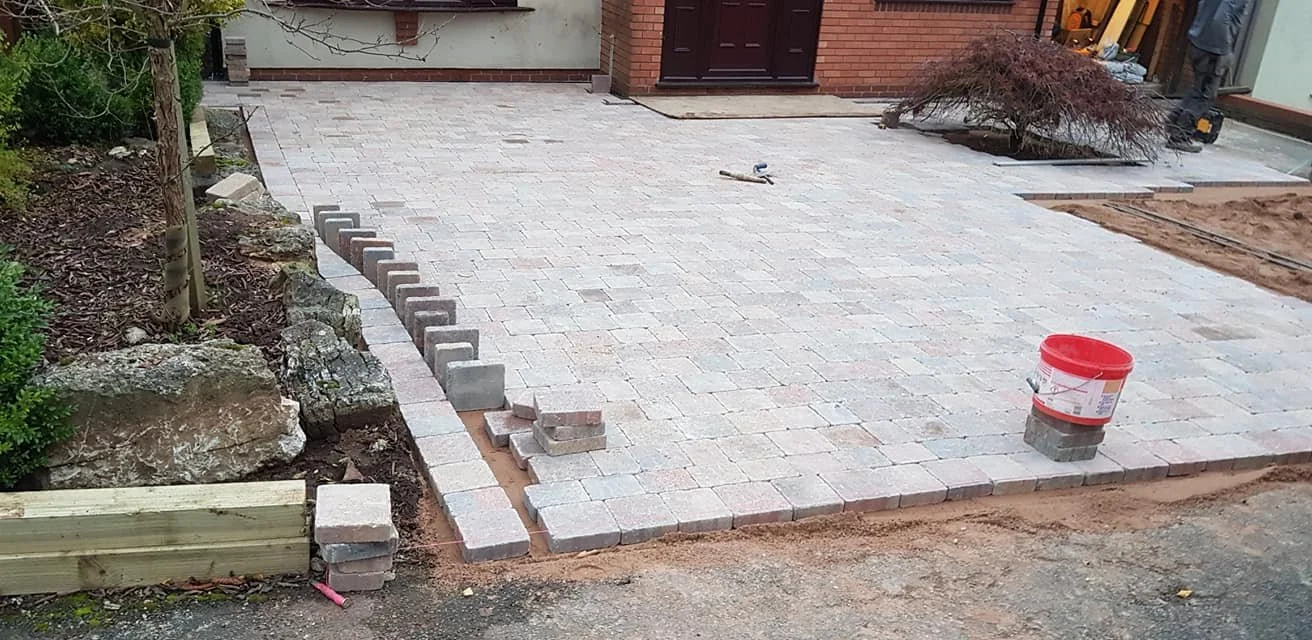Paved driveway under construction with bricks and pavers, a red bucket, and assorted tools. Part of the driveway is completed, with an edge of higher brick border on the left, and some bricks are stacked nearby. There's a mulch bed with rocks and a s