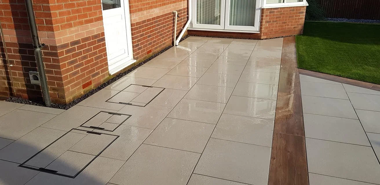 Clean, wet outdoor patio with beige tiles, a brick house wall, a white door, a white sliding door, and a strip of artificial grass.