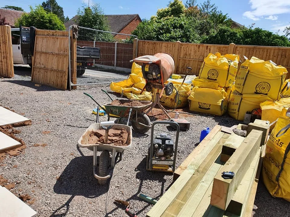 Construction site with wheelbarrows, bags of building materials, wooden planks, and construction equipment enclosed by a partially built wooden fence.