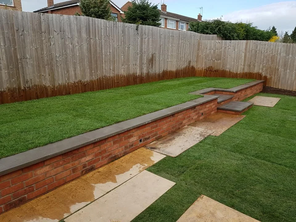 Backyard with a brick retaining wall, new grass, and outdoor steps with concrete slabs leading up to the elevated area.