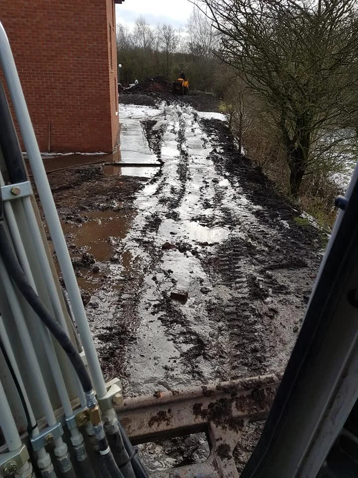View from inside a construction vehicle looking out at a muddy, uneven dirt path next to a brick building with a person operating a small yellow excavator at the top of the hill. The area appears wet and muddy, with some puddles.