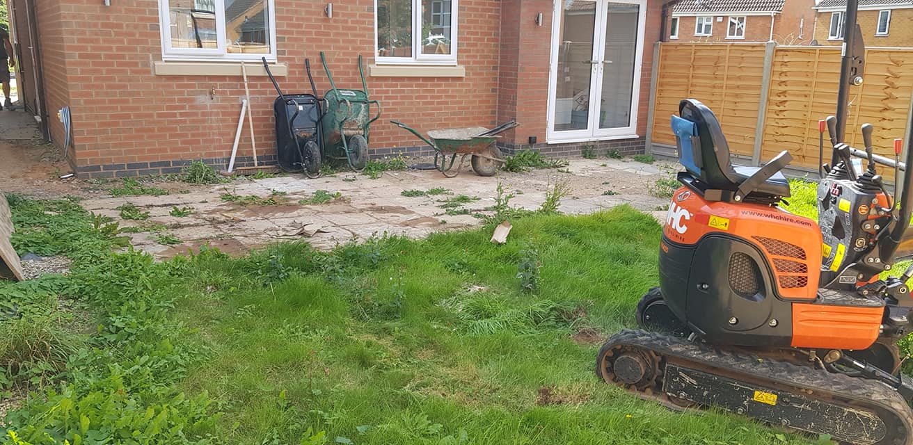 Backyard with construction equipment, including an orange mini excavator and wheelbarrows, and a partially paved area in front of a brick house with sliding glass doors.