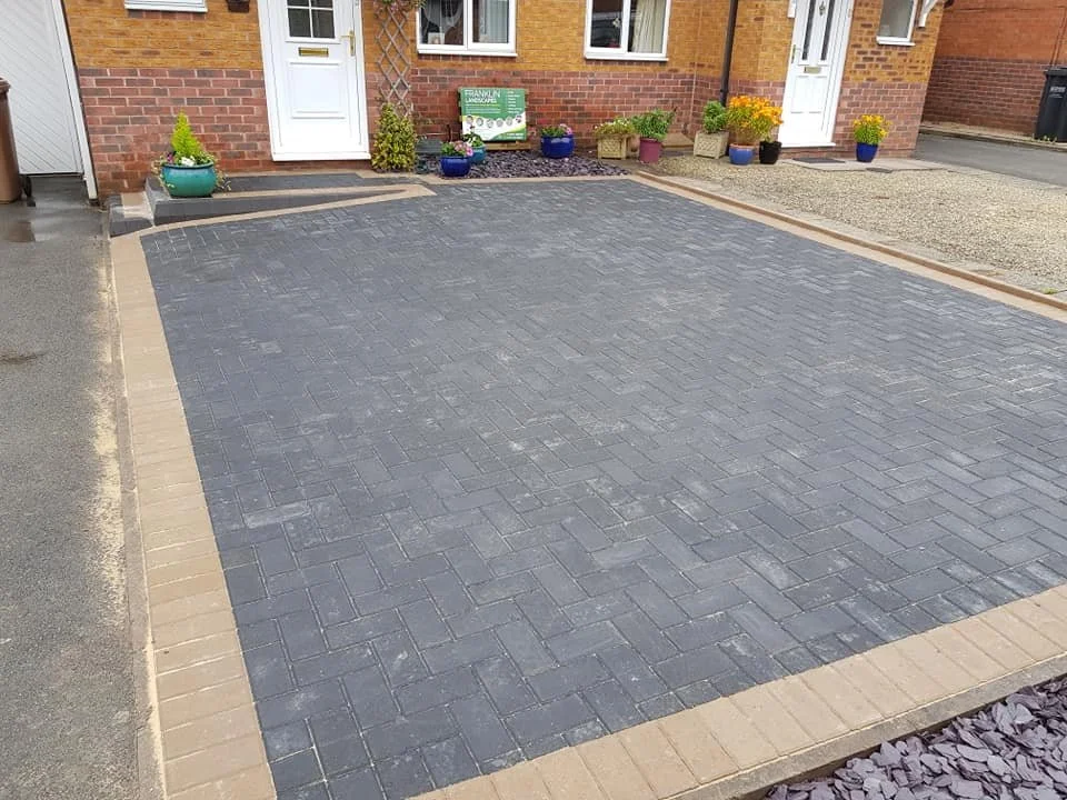 Freshly paved driveway with gray bricks bordered by beige bricks in front of a brick house, decorated with various potted plants near the entrance.