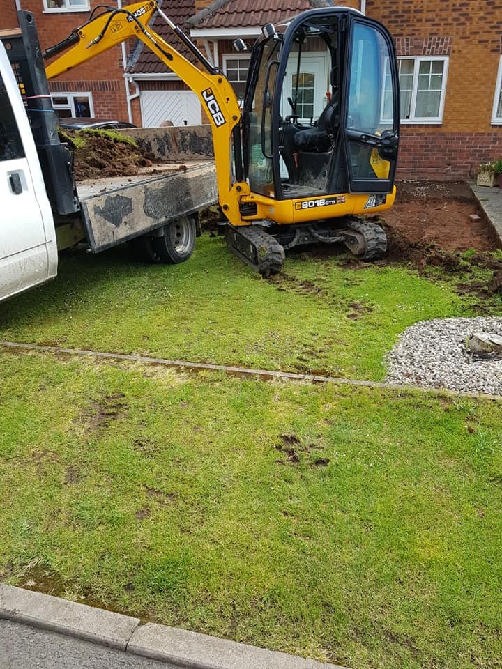 A small yellow excavator digging in a front yard near a sidewalk, with a truck parked nearby. The yard has some patches of disturbed grass and a gravel area.