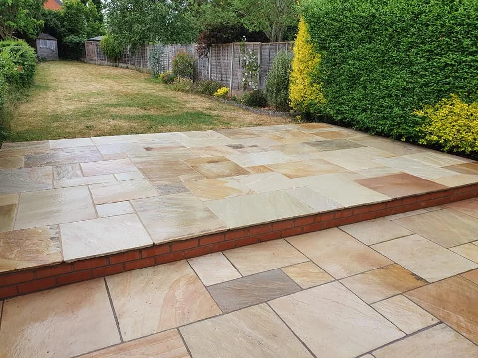 A backyard patio with beige stone tiles and a small brick wall at the edge. In the background, there is a grassy lawn with shrubs, trees, and a wooden fence.