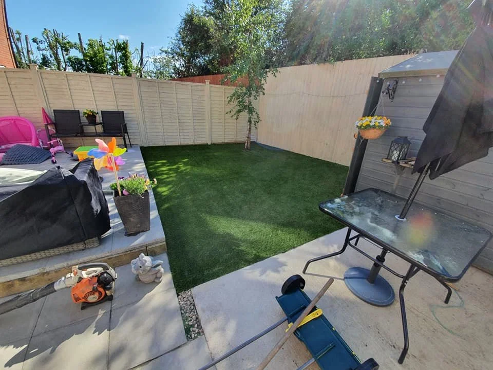Backyard with a grassy lawn, a fenced perimeter, a small tree, patio area with potted plants, outdoor furniture, colorful pinwheels, a table with an umbrella, and gardening tools.