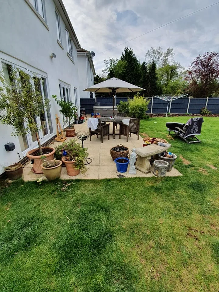 Backyard patio with potted plants, outdoor table with chairs and umbrella, a lounge chair, and various garden tools and decorations, next to a white house with sliding glass doors, a green lawn, and a fenced yard with trees and bushes.