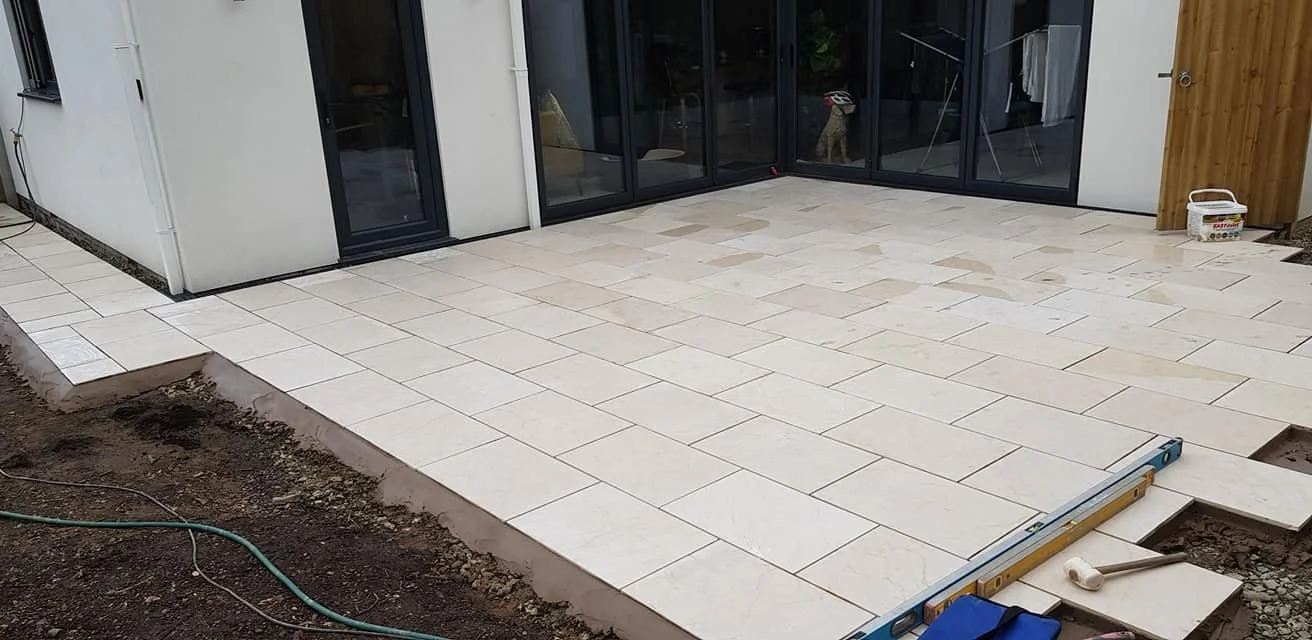 Construction site with a patio made of large beige tiles, adjacent to a house with glass sliding doors, and tools and materials for outdoor flooring.