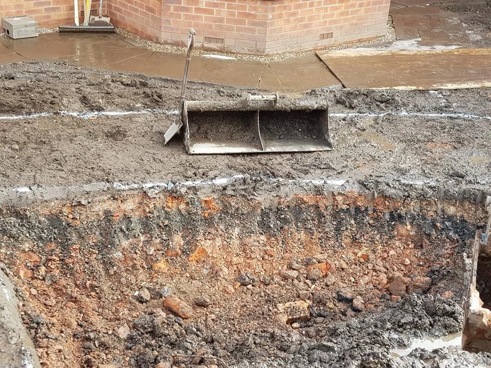 Construction site with excavated soil, a large rectangular pipe, and a partly poured concrete sidewalk near a brick building.