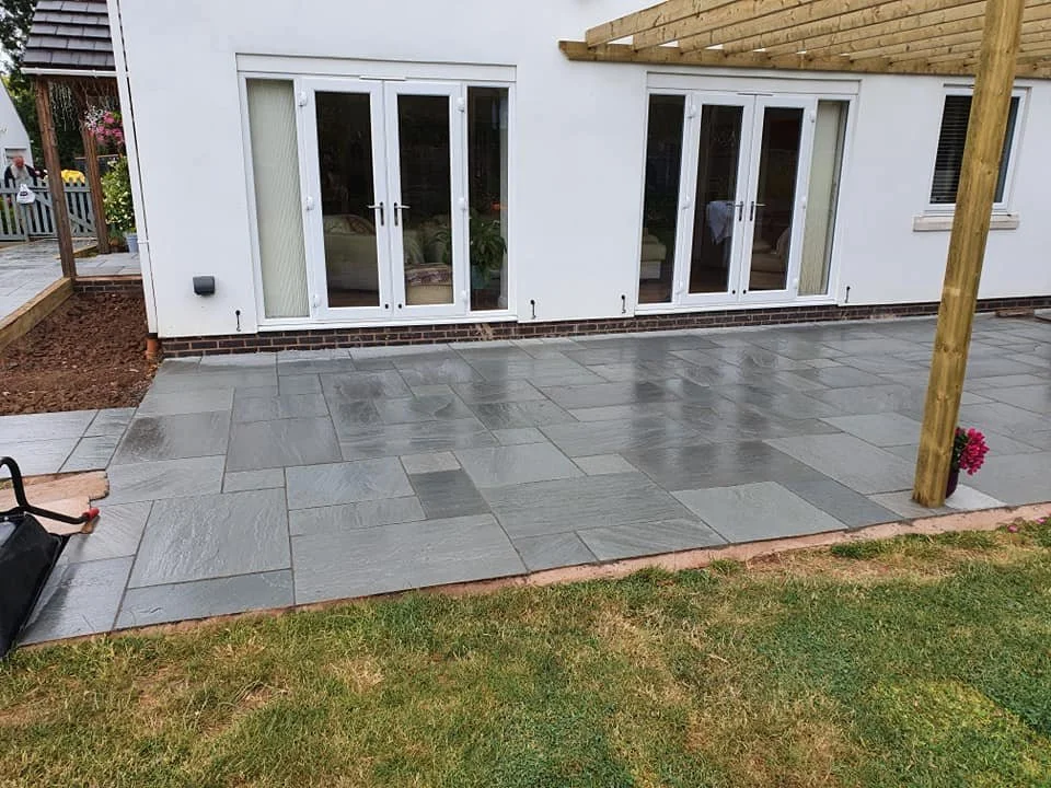 Recently installed grey stone patio in backyard with white house, large glass doors, wooden pergola frame, small garden bed, and grassy lawn area.