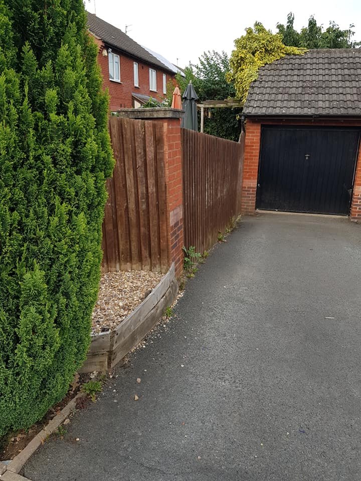 A corner of a driveway with a tall green hedge on the left, a wooden fence, and a brick wall with a garage door on the right.