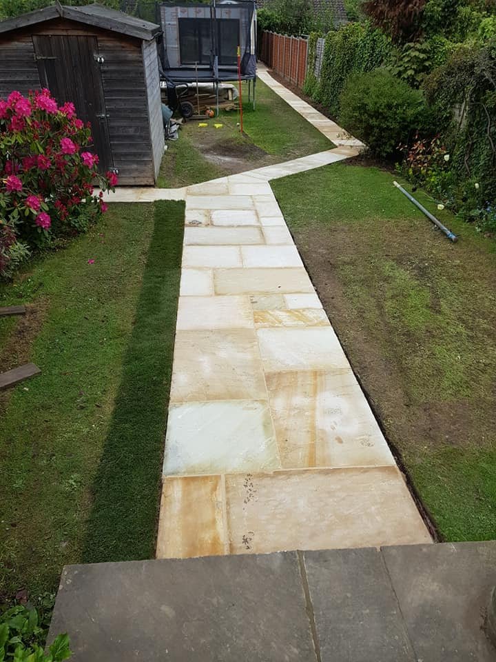 A backyard pathway made of large beige stone slabs, bordered by green grass and garden beds, leading to a small shed and trampoline.