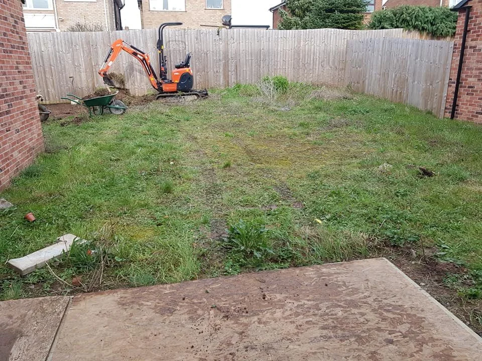 Backyard with a small orange excavator, a wheelbarrow, and patches of disturbed grass surrounded by a wooden fence and brick walls, indicating landscaping work.