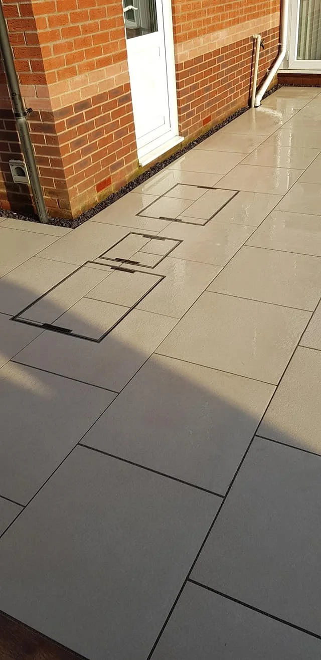 Three access panels on a light-colored tiled patio next to a brick wall and a door, with a shadow cast on the tiles.
