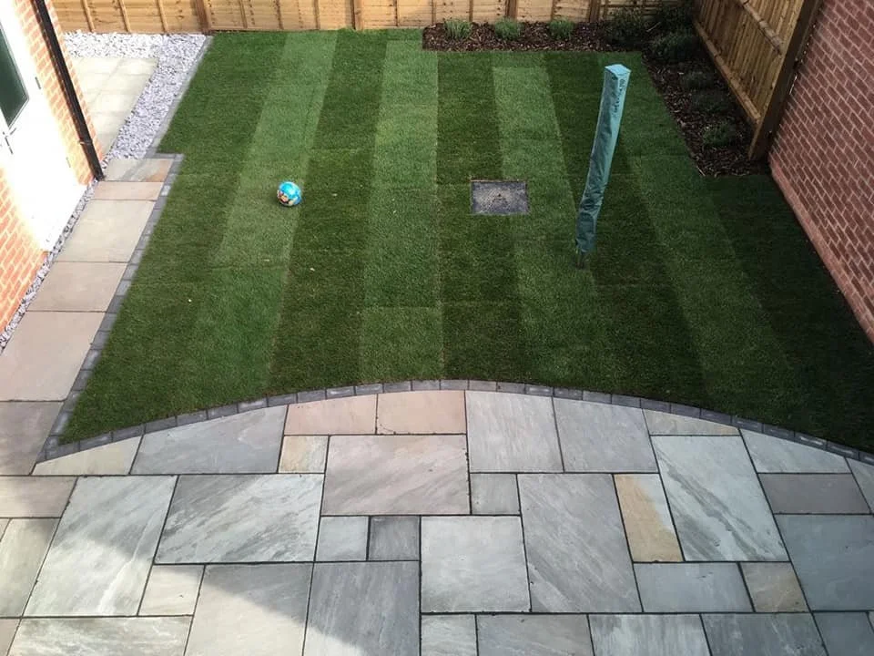 A backyard with a striped lawn, a small blue ball, a green garden post, and a patch of planting bed along the fence, enclosed by a wooden fence and brick walls, with a stone paved patio area in the foreground.