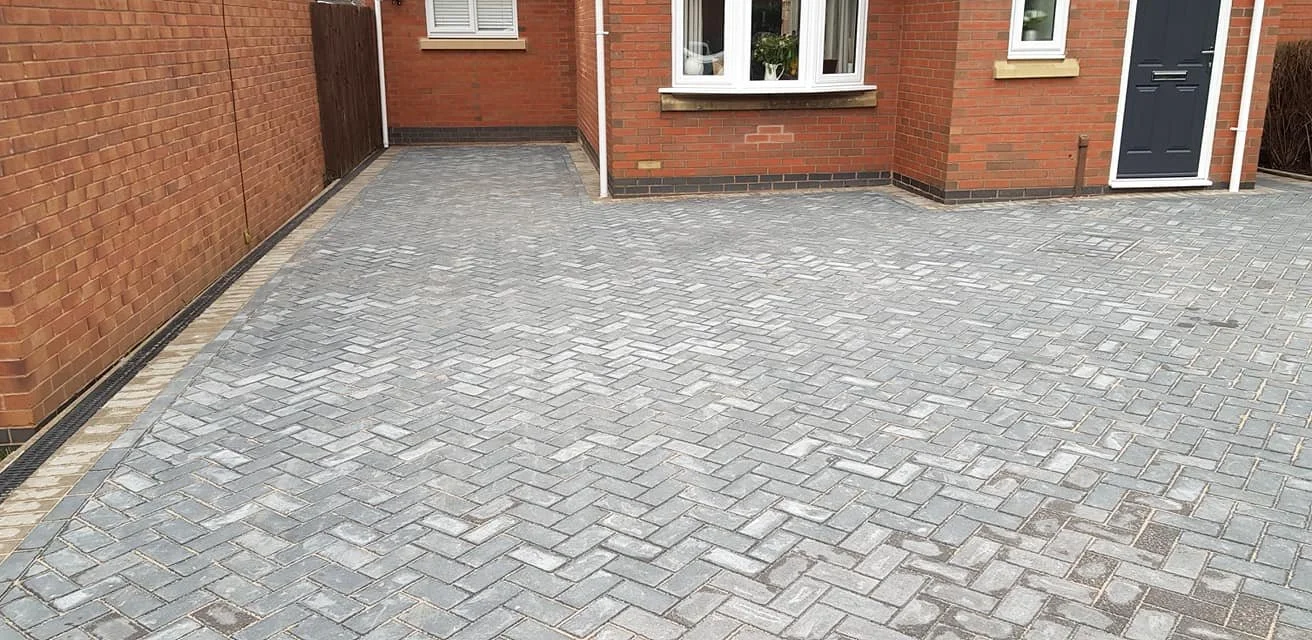 A paved driveway with grey interlocking bricks and a brick house with a blue door and white window frames.